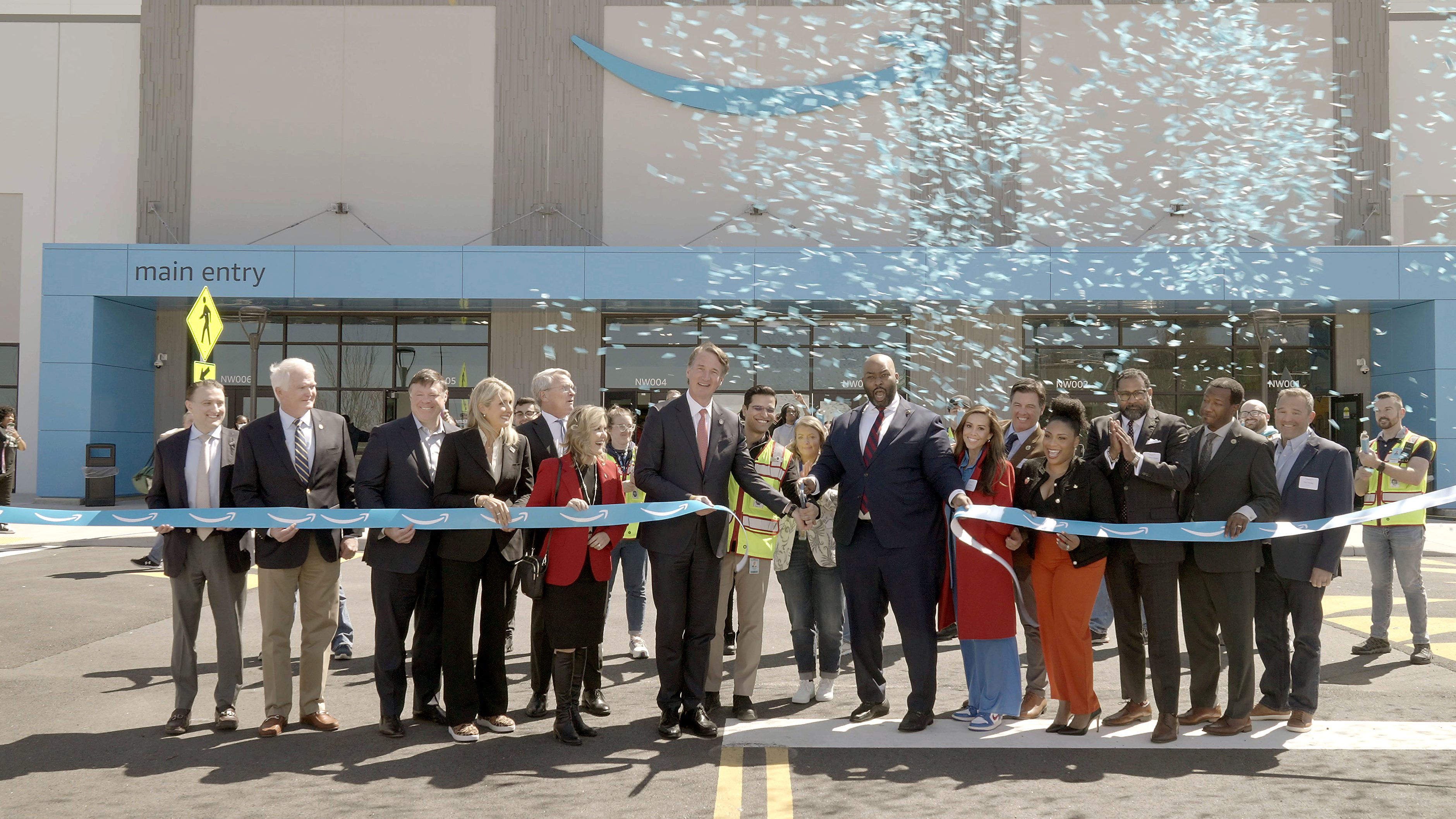 A ribbon-cutting ceremony at an Amazon facility featuring a group of people in business attire standing in front of the main entrance. Confetti is released as two individuals cut the blue ribbon with Amazon logos.