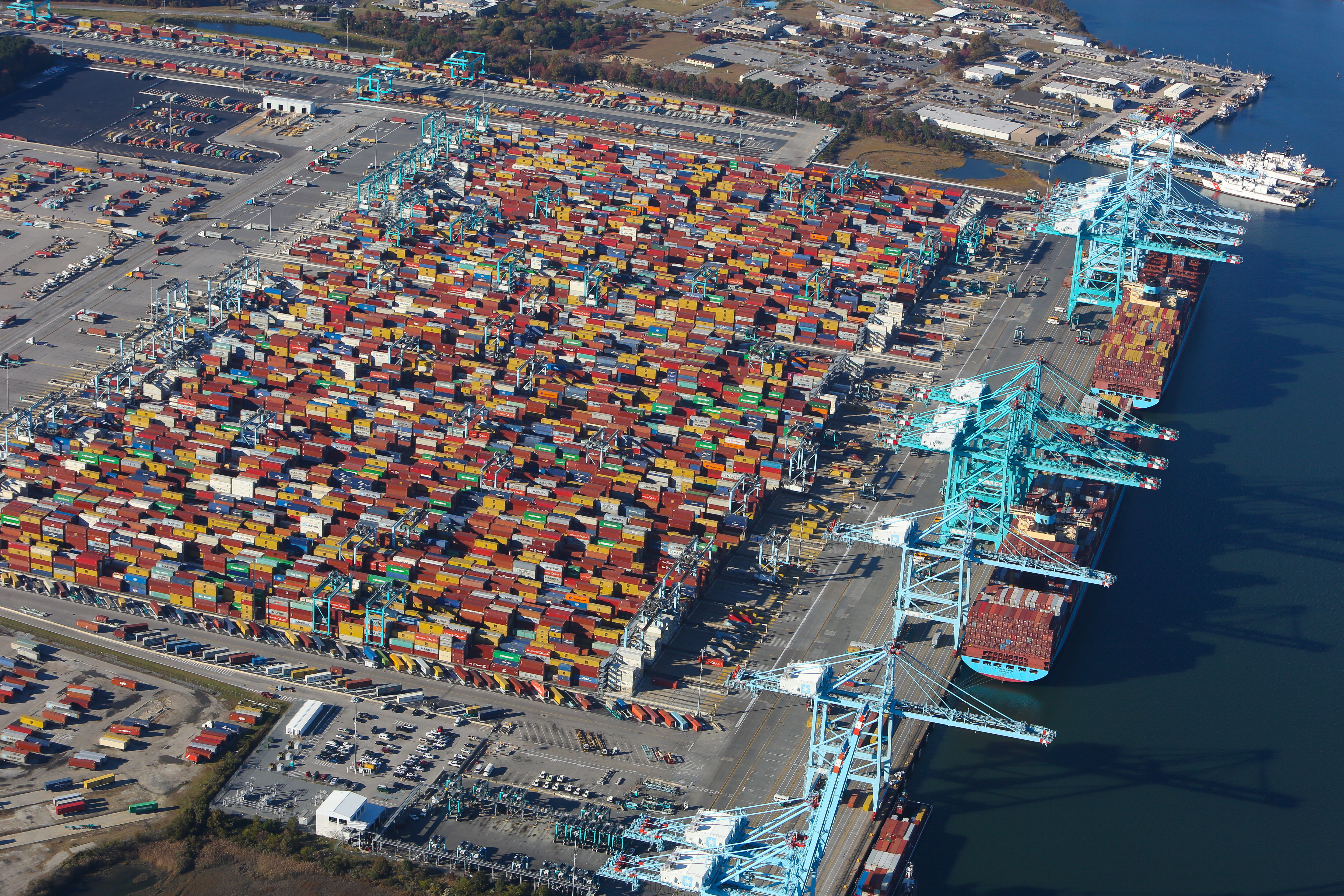 Aerial view of a busy shipping port with numerous colorful shipping containers stacked neatly in rows and large blue cranes along the waterline.